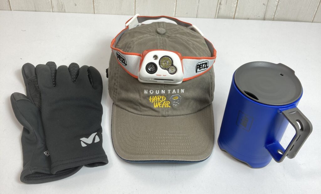 黒いアウトドアグローブ、ヘッドランプ付きのMOUNTAIN HARDWEARキャップ、青い携帯用マグカップが机に並べられている、登山やアウトドア活動に必要な小物を示す画像です。